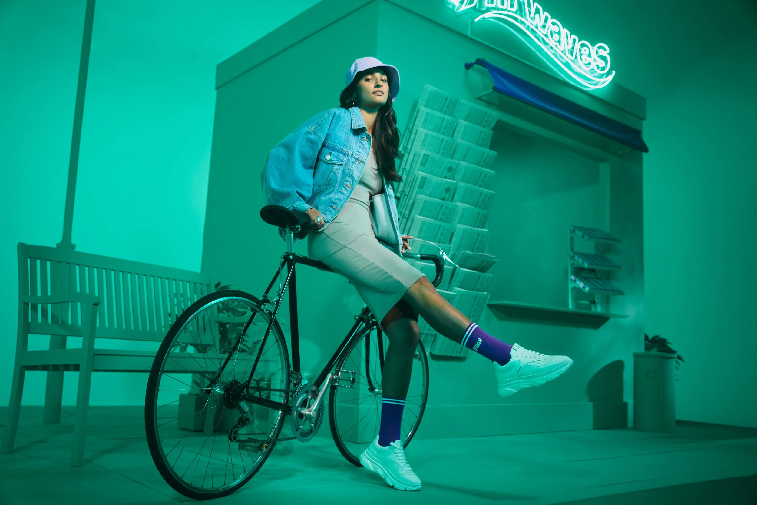 Eine junge Frau mit Bucket Hat auf einem Rennrad vor dem Kiosk, an den Füßen lila Airwaves-Socken