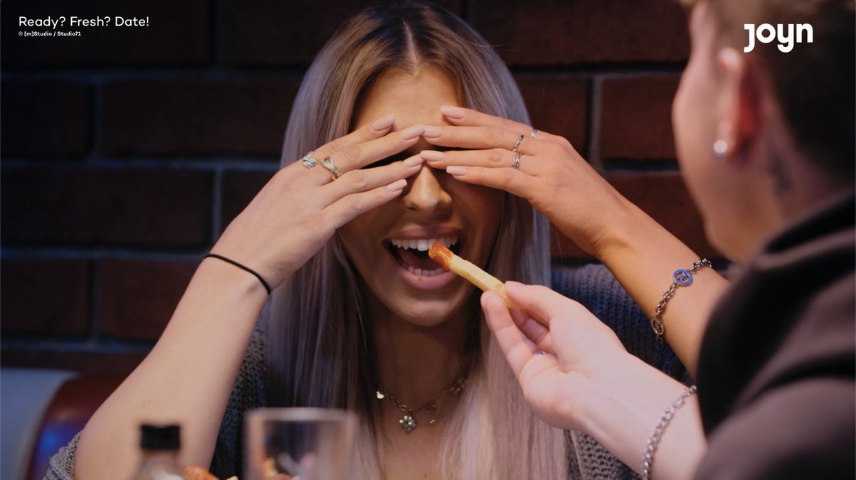 Eine Frau mit verbundenen Augen bekommt von ihrem Date eine Pommes gefüttert