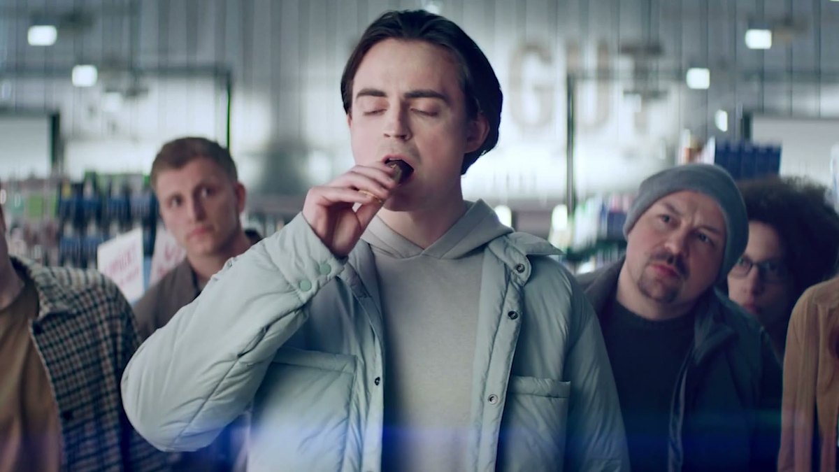 Ein junger Mann beißt mit geschlossenen Augen in einen Wunderbar-Riegel, umringt von anderen Kunden